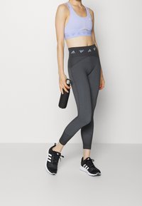 Purple sports bra and black textured leggings with a high waistband, paired with black athletic shoes featuring white accents, holding a water bottle.