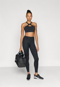 Μαύρο αθλητικό σετ με κορυφή αθλητικού σουτιέν με κοψίματα και κολάν με ψηλή μέση. Μαύρα αθλητικά παπούτσια και τσαντάκι duffle που ταιριάζει.