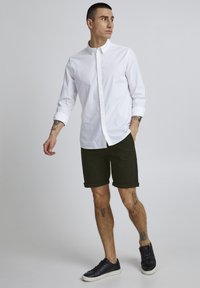 Chemise blanche à boutons, shorts verts et baskets foncées. La chemise est à manches longues ; les shorts sont à longueur genou avec des ourlets roulés.