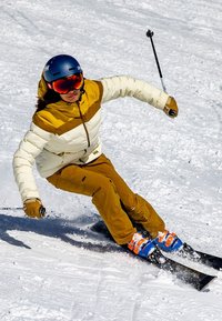 Skikleding met een witte en bruine geïsoleerde jas, bruine broek, blauwe en oranje skischuilen, en een blauwe helm met spiegelende goggles op de sneeuw.