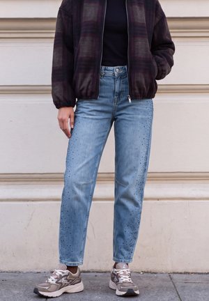 Une personne porte un jean bleu clair orné, une veste foncée zippée et des baskets épaisses devant un mur en pierre claire.