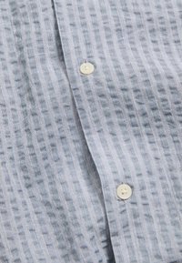 Camicia azzurra a righe con bottone, realizzata in un tessuto texturizzato, presenta due bottoni bianchi visibili lungo la boutonnière, mostrando un design rilassato e arioso.