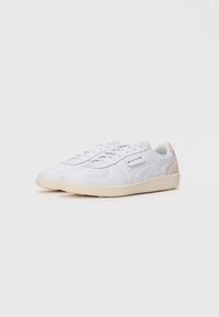 Et par hvite sneakers med lav topp og beige såler, samt semsket hæl-akenter, plassert side om side på en enkel lys bakgrunn.