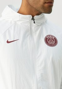 Valkoinen Nike-takki, jossa on vetoketju edessä, punainen "PARIS" -logo ja tyylitelty emblem, sileä pinta ja huppu.