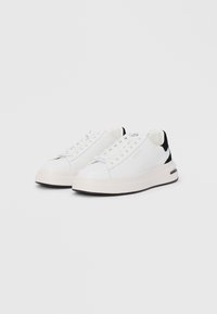 Baskets blanches avec une tige en cuir lisse, un accent noir au niveau du talon et une semelle intermédiaire blanche épaisse. Elles présentent des lacets plats et un branding discret.