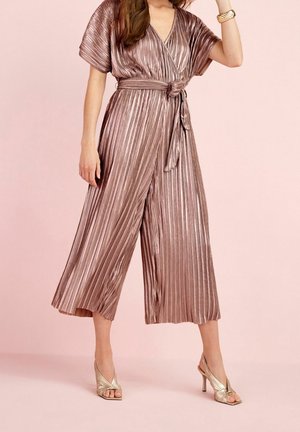 Vrouw draagt een geplooide metallic roze gouden jumpsuit met korte mouwen, een gestrikte taille en gouden sandalen met hoge hakken, tegen een roze achtergrond.