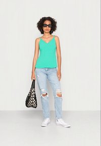 Camisola texturizada em turquesa com alças finas, combinada com jeans rasgados em azul claro e ténis brancos. Modelo segura uma bolsa de rede preta.