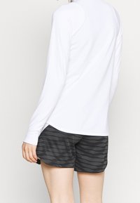 Witte top met lange mouwen en een ronde hals, gecombineerd met zwarte shorts met een golvend patroon, die lichte en ademende materialen benadrukken.