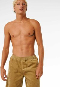 Beige Cordhosen mit elastischem Bund, getragen von einem oberkörperfreien männlichen Model. Auffälliges Tattoo auf der linken Brustseite.