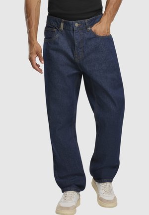 Mann iført løse mørkeblå denimjeans med hendene i lommene og hvite joggesko, stående mot en ensfarget lys bakgrunn.
