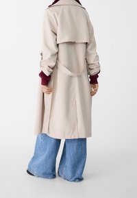 Beige trenchcoat med dubbelknäppt design, bälte i midjan och uppvikbar krage, kombinerad med vida ljusblå jeans.