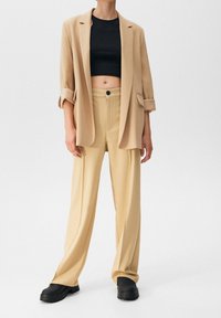 Blazer beige surdimensionné sur un crop top noir, associé à un pantalon beige taille haute et des chaussures noires robustes. Textures lisses, détails minimalistes.