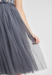 Robe en tulle gris à texture à pois, taille marquée et broderie florale sur le corsage, avec une jupe fluide et superposée.