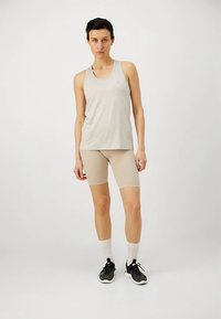 Person steht und trägt ein beiges ärmelloses Oberteil, passende enge Shorts, weiße Socken und schwarze Sportschuhe vor einem schlichten weißen Hintergrund.