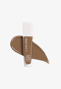 Fond de teint Lancôme dans un flacon en verre transparent avec un capuchon blanc, affichant un liquide marron moyen lisse avec une finition brillante.