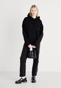 Felpa con cappuccio nera e tasca frontale, abbinata a jeans scuri. Gli accessori includono una piccola borsa nera e scarpe lucide nere con dettagli rossi.