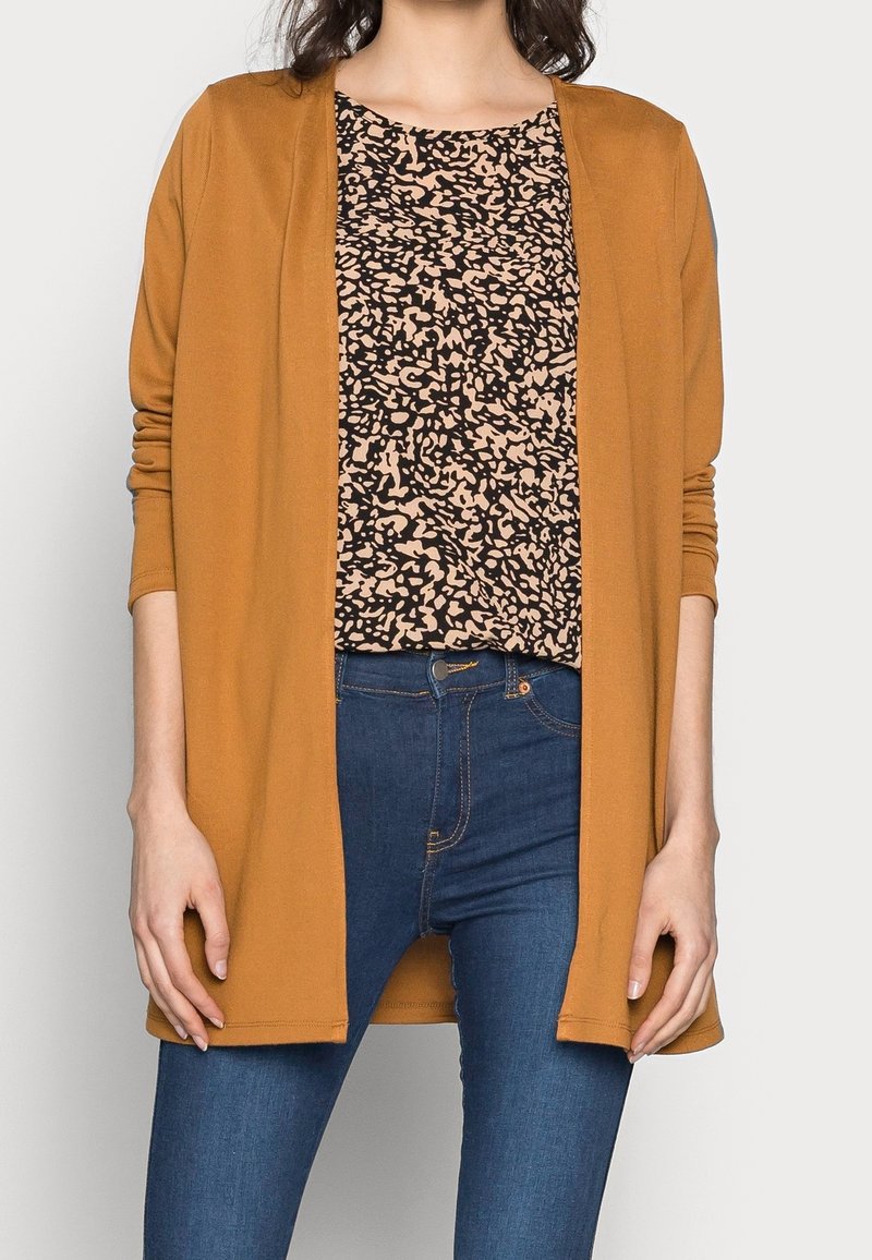 Cardigan jaune moutarde porté sur un haut à motifs marron et noir, associé à un jean bleu ajusté. Le cardigan a une coupe décontractée et des manches longues.