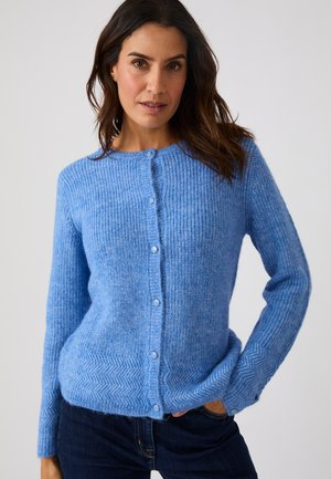 Femme portant un cardigan bleu tricoté boutonné avec des motifs en chevrons sur les poignets et l'ourlet, associé à un jean bleu foncé sur un fond uni.