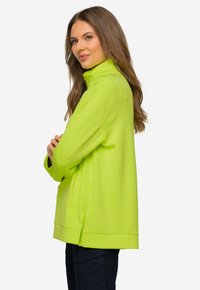 Sudadera de color verde neón con cuello alto, mangas largas y bolsillos laterales; textura suave y corte holgado con un dobladillo caído en la parte posterior.