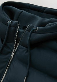 Hoodie rembourré bleu foncé avec fermeture éclair, matériel en métal argenté, et capuche ajustable avec cordons, présentant une texture lisse.