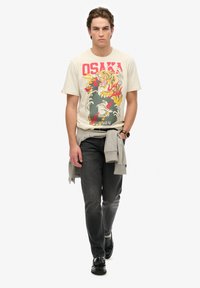 Camiseta de algodón beige con un gráfico de tigre colorido, combinada con jeans gris oscuro y zapatos negros, con un sudadera gris atada a la cintura.