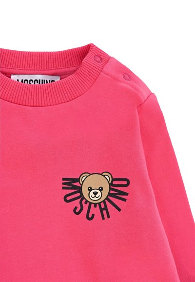 Felpa rosa con scollo a costine, caratterizzata da una grafica di un orso e il testo "MOSCHINO". Il materiale sembra morbido, con chiusure a pressione sulla spalla.
