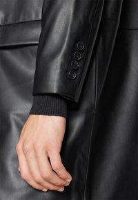 Rękaw czarnej skórzanej kurtki z prążkowanym czarnym mankietem, ozdobiony czterema guzikami wzdłuż boku. Gładka faktura z dopasowanym krojem.