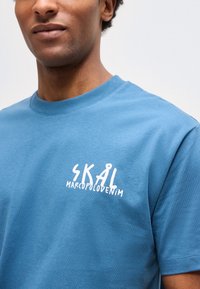 Άνδρας που φορά μπλε T-shirt με το λευκό τύπωμα «SKÅL MARCOPOLO DENIM» στο αριστερό στήθος, μπροστά σε απλό φωτεινό φόντο.