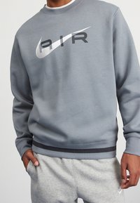 Šedá mikina s kulatým výstřihem, s velkým černým a stříbrným logem Nike, žebrovanými manžetami a tmavým akcentem na lemu.