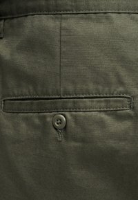 Ύφασμα σε ελιά πράσινο με λεία υφή, διαθέτει τσέπη με κουμπί και ραφές που έχουν ραφτεί με ακρίβεια. Το κουμπί είναι στρογγυλό με τέσσερις τρύπες.