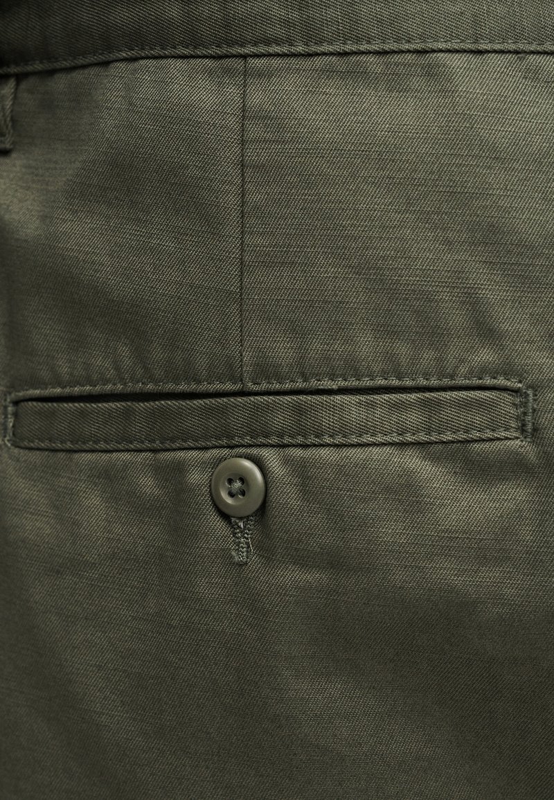 Tissu vert olive à la texture lisse, doté d'une poche à bouton et de détails de couture ajustés. Le bouton est rond avec quatre trous.