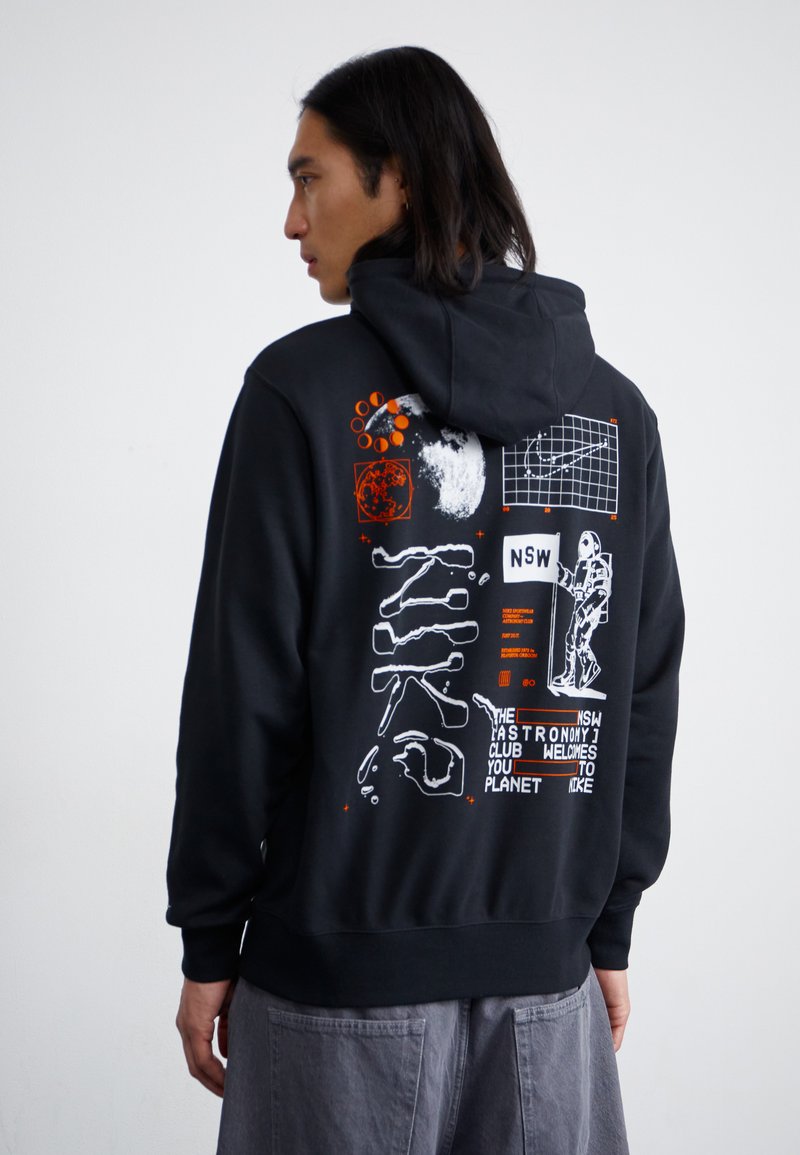 Un hoodie noir avec un design graphique au dos, présentant des illustrations, du texte et des motifs blancs et oranges liés à l'astronomie.