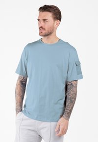 Mann mit Ärmeltattoos, der ein hellblaues kurzärmliges T-Shirt mit einer Tasche am linken Arm und hellen Hosen trägt, und nach links schaut.