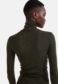 Personne avec les cheveux courts portant un col roulé ajusté de couleur vert foncé, tourné sur le côté, contre un fond clair et uni.