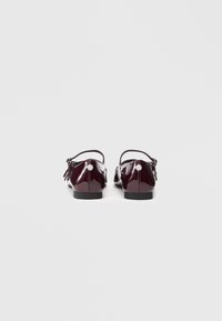 Tumši sarkani lakoti ādas sieviešu slingback dzīvok�ļi ar potīšu siksnām un sudraba apaļajiem logotipa elementiem uz papēžiem.