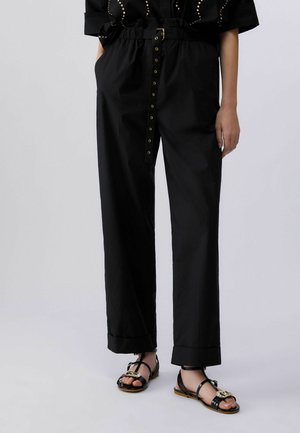 Frau trägt schwarze, weit geschnittene Hose mit hohem Bund und Gürtel sowie schwarze Sandalen mit goldenen Schnallendetails.