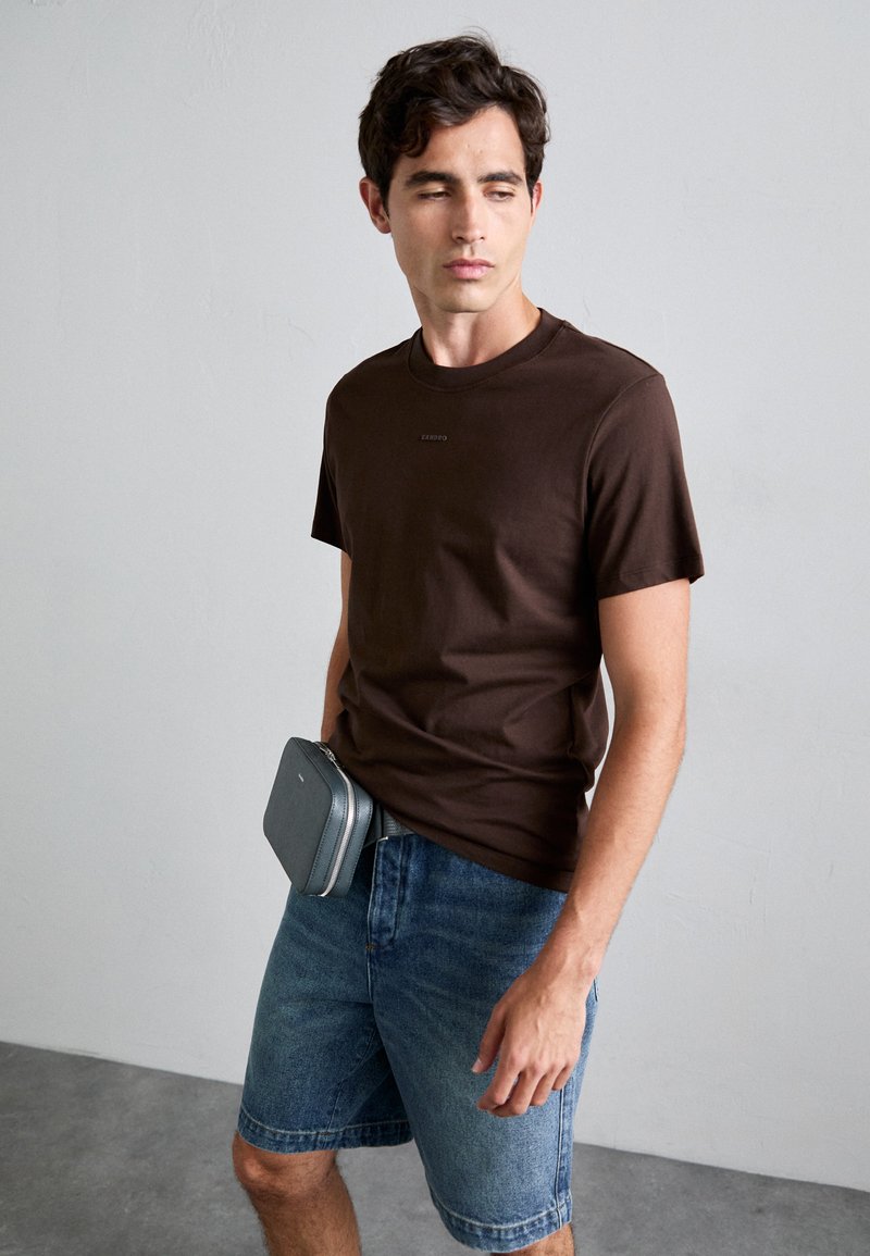 Maglietta in cotone marrone a maniche corte, abbinata a pantaloni shorts in denim blu. Una pochette rettangolare grigia è indossata in vita, fissata con una cintura.