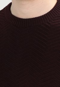 Burgundy stickad tröja som har ett texturerat fiskbensmönster och en ribbad rund halsringning. Nära bild av tyg- och stickdetaljer.