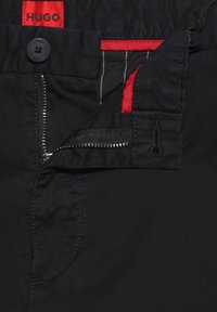 Pantalon noir avec fermeture éclair métallique partiellement ouverte, bouton noir et étiquette de marque rouge indiquant Hugo à l'intérieur de la ceinture.