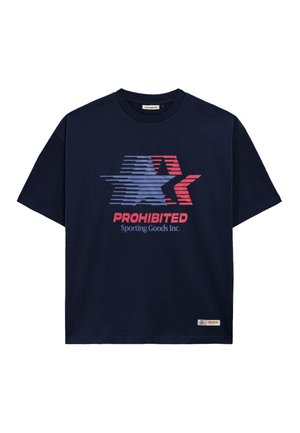 Ung mand iført navy kasket og navy T-shirt med rød, hvid og blå stjerne grafik og teksten "PROHIBITED Sporting Goods Inc."