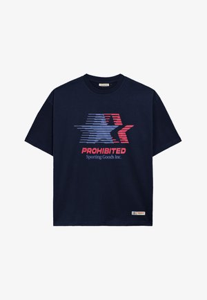 Giovane uomo che indossa un cappello navy e una T-shirt navy con grafica di stella rossa, bianca e blu e testo "PROHIBITED Sporting Goods Inc."