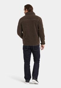 Ruskea fleece-takki, jossa on korkea kaulus, teksturoitu pinta ja takasaumadetaili, yhdistettynä tummiin farkkusiin ja mustiin lenkkareihin.