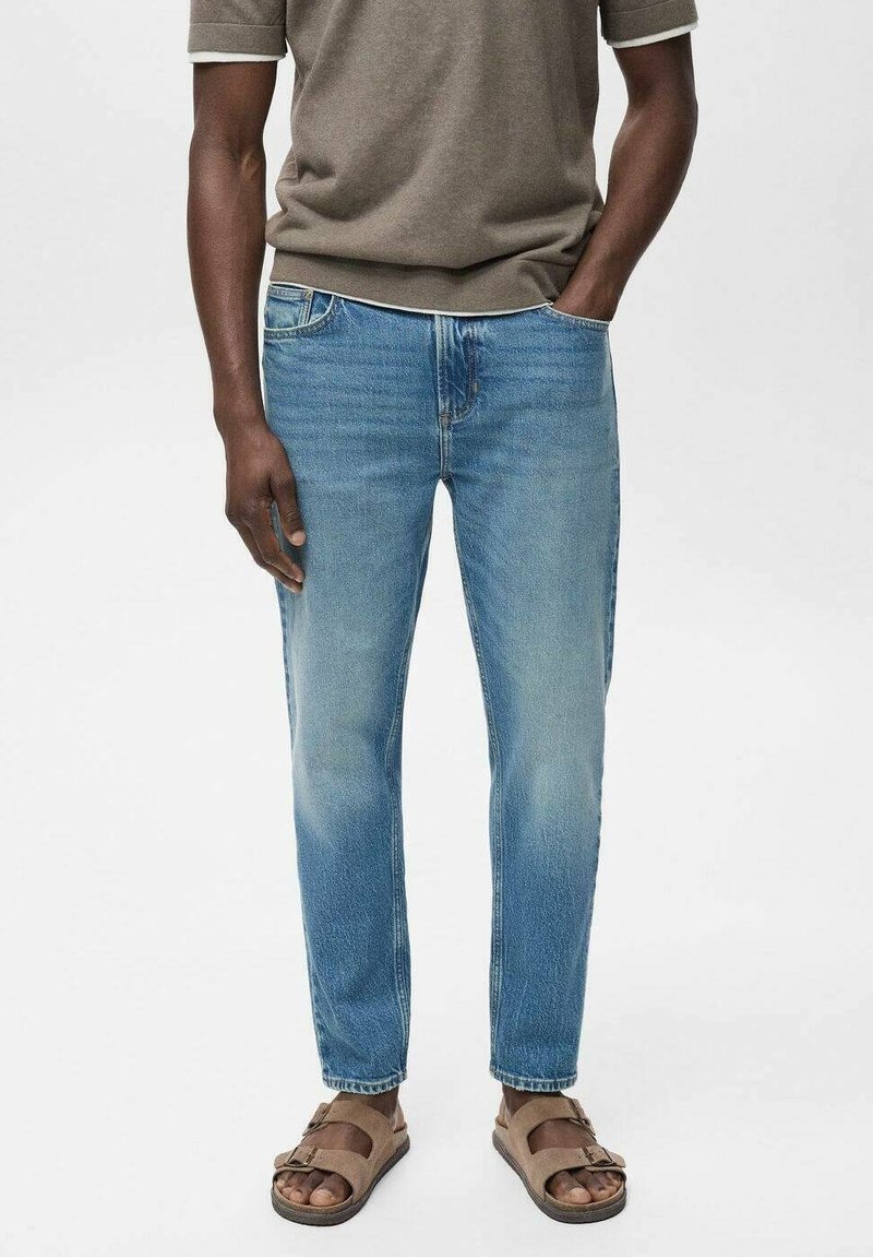Homme portant un jean bleu clair, une chemise marron à manches courtes et des sandales ouvertes marron, debout avec une main dans la poche.