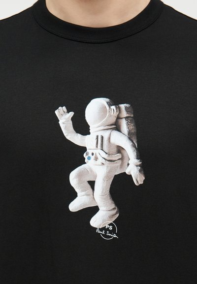 Άστροναύτης σε διαστημική στολή με σακίδιο πλάτης, με το δεξί χέρι σηκωμένο, τυπωμένος με λευκά γράμματα σε μαύρο ύφασμα, με μικρό λογότυπο "PS Paul Smith" από κάτω.