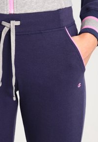 Gros plan sur un pantalon de jogging bleu marine avec des bordures roses et un logo, un cordon gris, et une main glissée dans la poche, porté avec une veste assortie.