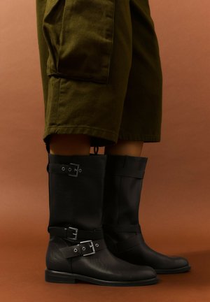 Bottes en cuir noires mi-mollet avec deux boucles portées avec des shorts cargo vert olive longueur genou sur fond marron.