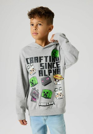 Ragazzo che indossa una felpa con cappuccio Minecraft grigio chiaro con la scritta "Crafting Since Alpha" e teste di personaggi pixelate, abbinata a jeans blu chiaro.