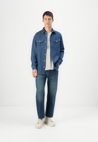 Chaqueta de mezclilla de color azul oscuro con dos bolsillos en el pecho, llevada sobre una camiseta de color gris claro. Combinada con jeans rectos de color azul medio y zapatillas deportivas blancas.