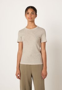 Young woman with curly hair wearing beige short-sleeve top and olive green pants, standing against a plain light background.