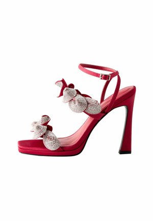 Czerwone satynowe sandały na wysokim obcasie mają duży, ozdobny kokardę z biżuterią, cienkie paski wokół kostki oraz błyszcząca czarna podeszwę. Obcas jest ostro kątowany.